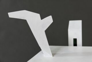Abstrakte weiße geometrische Skulptur mit einer kantigen Form, die sich von einem flachen Sockel nach oben erstreckt; ein rechteckiger Block mit einem kleinen vertikalen Schlitz steht im Hintergrund vor einem dunklen Hintergrund.