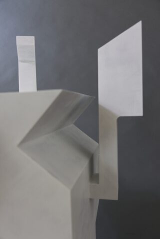 Abstrakte geometrische Skulptur mit scharfen Winkeln und flachen Oberflächen aus hellgrauem Material, vor einem dunklen, neutralen Hintergrund.