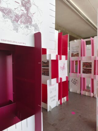 Ausstellungsinstallation aus modularen roten, rosa und weißen Paneelen mit Architekturzeichnungen und Karten, angeordnet in einem Galerieraum mit Betonboden.