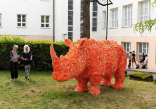 Große orangefarbene Nashornskulptur aus Blättern, die auf einer Rasenfläche in der Nähe eines Gebäudes ausgestellt ist, während Menschen in der Nähe spazieren gehen und sie betrachten.