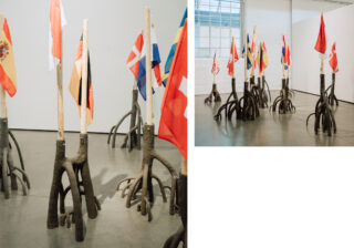 Mehrere Nationalflaggen sind an Holzstangen mit geschnitzten, wurzelartigen Sockeln befestigt und in einem minimalistisch gestalteten Galerieraum mit weißen Wänden und Betonböden angeordnet.