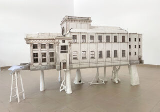 In einem Galerieraum steht ein detailliertes Architekturmodell eines neoklassizistischen Gebäudes auf weißen, baumartigen Stützen. Links davon steht ein kleiner weißer Hocker mit Papieren darauf.