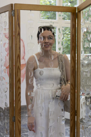 Eine Frau in einem weißen Kleid steht hinter einem hölzernen Raumteiler, der mit hängenden Brillenfassungen verziert ist.