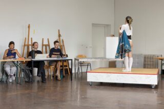 Drei Schüler sitzen an ihren Schreibtischen und zeichnen eine Figur, die auf einer Plattform steht.