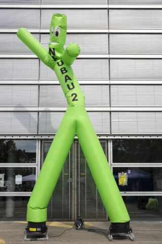 Vor einem modernen Gebäude mit horizontalen Metalljalousien steht eine grüne aufblasbare Figur, auf deren Seite „NEUBAU 2“ aufgedruckt ist.