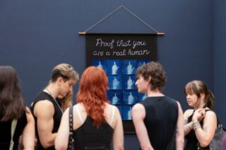 Die Menschen stehen vor einem Wandbehang, auf dem über einem Raster aus blauen Bildern, die einen ausgestreckten Mittelfinger zeigen, die Aufschrift „Beweis, dass du ein echter Mensch bist“ zu lesen ist.