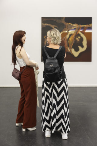 Zwei Frauen, die vor einem abstrakten Gemälde in einer Galerie stehen und lachen.