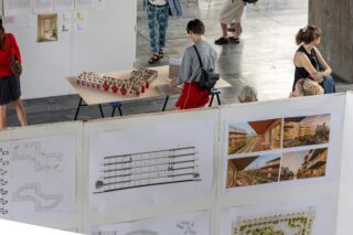 Ein Student betrachtet ein Architekturmodell auf einem Tisch vor Ausstellungstafeln, auf denen Baupläne und Visualisierungen zu sehen sind.
