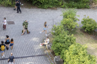 Menschen, die auf einem gepflasterten Weg in der Nähe von Büschen entlanggehen, wobei eine Person auf einem Steinblock sitzt.