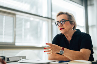 Eine Frau in schwarzer Kleidung und mit Brille gestikuliert, während sie bei einer Besprechung in einem hellen Büro spricht.