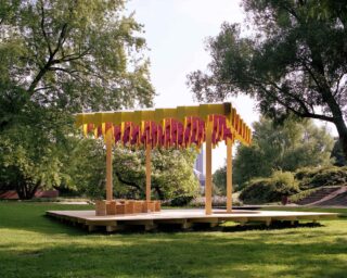 Ein Holzpavillon im Freien mit einem gelb-rosa verzierten Dachvorsprung, der auf einer niedrigen Terrasse in einem Park steht.