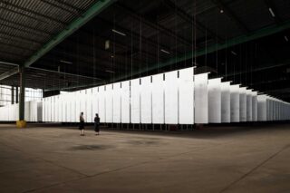 Zwei Personen stehen in einer großen Industriehalle und betrachten eine Kunstinstallation aus hängenden weißen Paneelen.