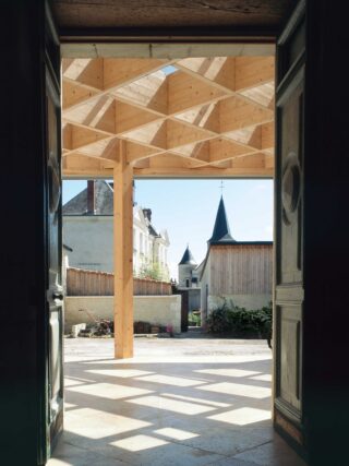 Blick durch eine offene Tür auf einen Innenhof mit einer geometrischen Dachkonstruktion aus Holz und in der Ferne sichtbaren Kirchtürmen.