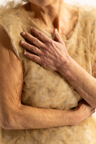 Nahaufnahme der Hand einer Person, die auf ihrer Brust ruht; sie trägt ein beiges, mehrlagiges Netzoberteil.