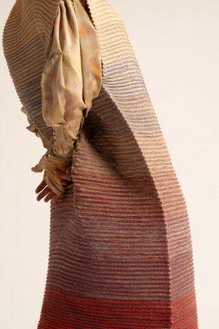 Eine Person, die ein plissiertes, mehrfarbiges Strickkleid über einem hellbraunen, im Tie-Dye-Stil gefärbten Langarm-Oberteil trägt und die Hände hinter dem Rücken verschränkt hat.