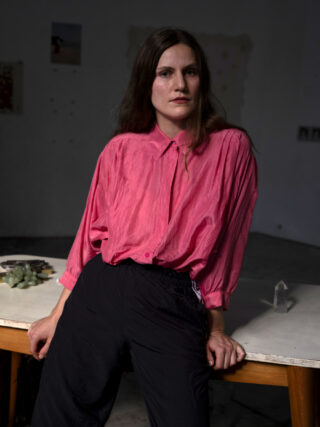Eine Frau, die ein leuchtend pinkfarbenes Hemd und eine schwarze Hose trägt, lehnt an einem Tisch.