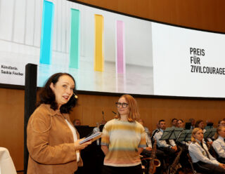Eine Frau spricht an einem Rednerpult, hinter ihr ist auf einer Leinwand die Aufschrift „Preis für Zivilcourage“ sowie der Name der Künstlerin Saskia Fischer zu sehen.