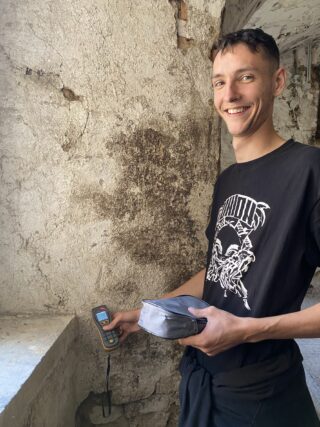Ein junger Mann, der lächelnd ein tragbares digitales Feuchtigkeitsmessgerät an einer strukturierten Wand einsetzt.
