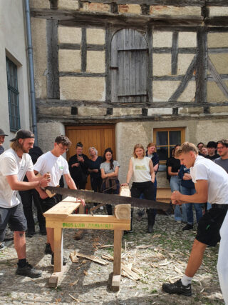 Zwei Personen zersägen mit einer großen Handsäge einen Baumstamm auf einer hölzernen Werkbank, während eine Gruppe von Menschen vor einem Fachwerkhaus zusieht.