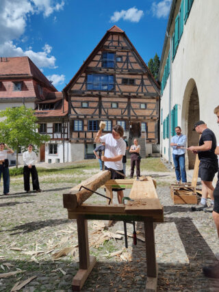Eine Person bearbeitet mit einem Holzhammer einen Holzbalken auf einer Werkbank, während andere ihr in einem Innenhof vor einem Fachwerkhaus zusehen.