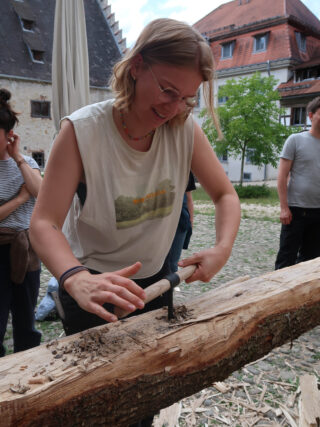 Eine Frau, die mit einem Ziehmesser Holz von einem Baumstamm abschneidet.
