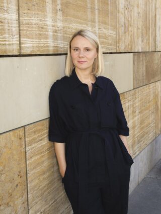 Eine Frau mit blonden Haaren, die einen dunkelblauen Overall mit Gürtel trägt, lehnt an einer Steinmauer.
