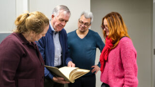 Vier Menschen stehen beieinander und blicken auf ein aufgeschlagenes Buch hinab.